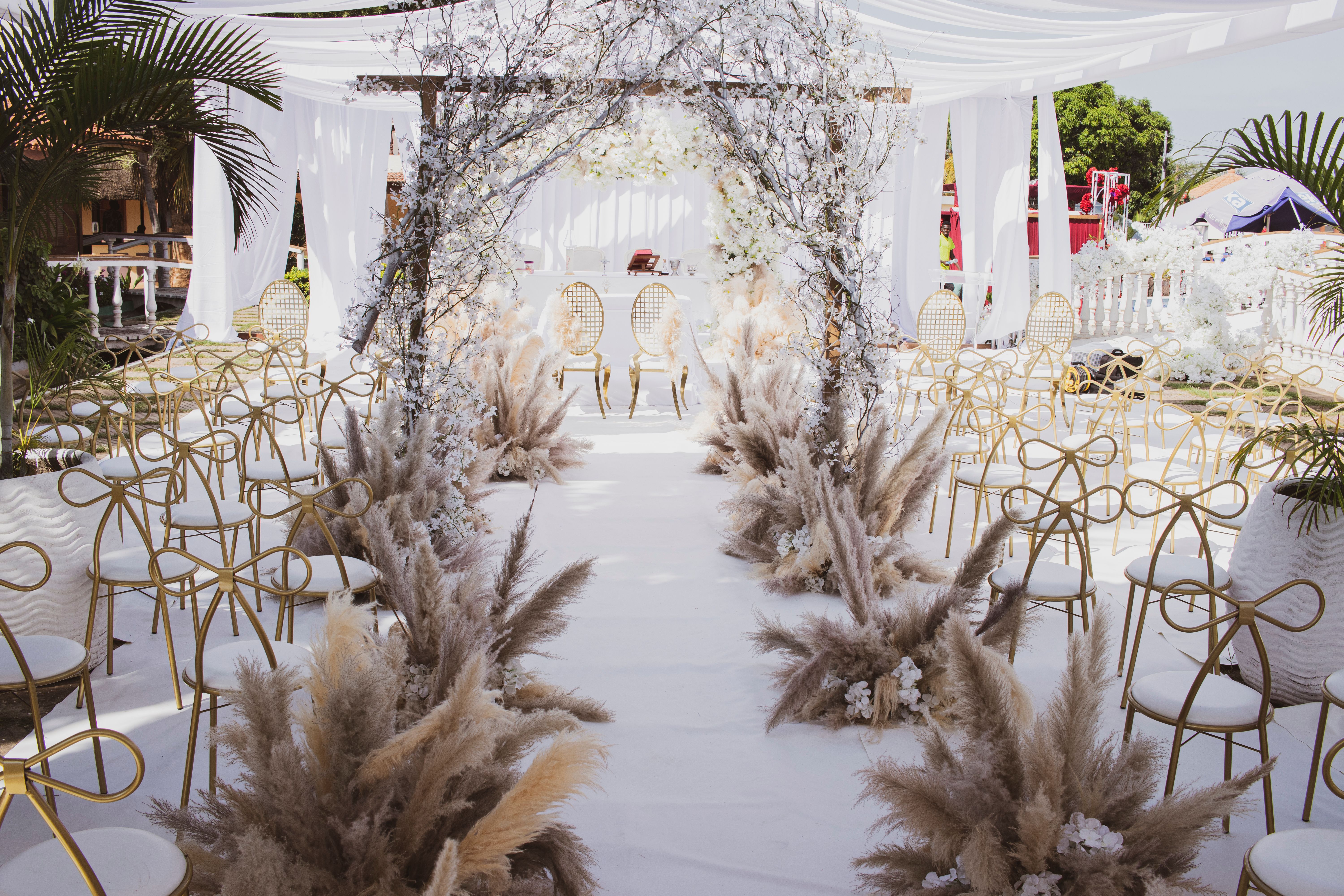 Elegant outdoor wedding setup in Angola featuring a white floral arch and golden chairs under a sunny sky.
