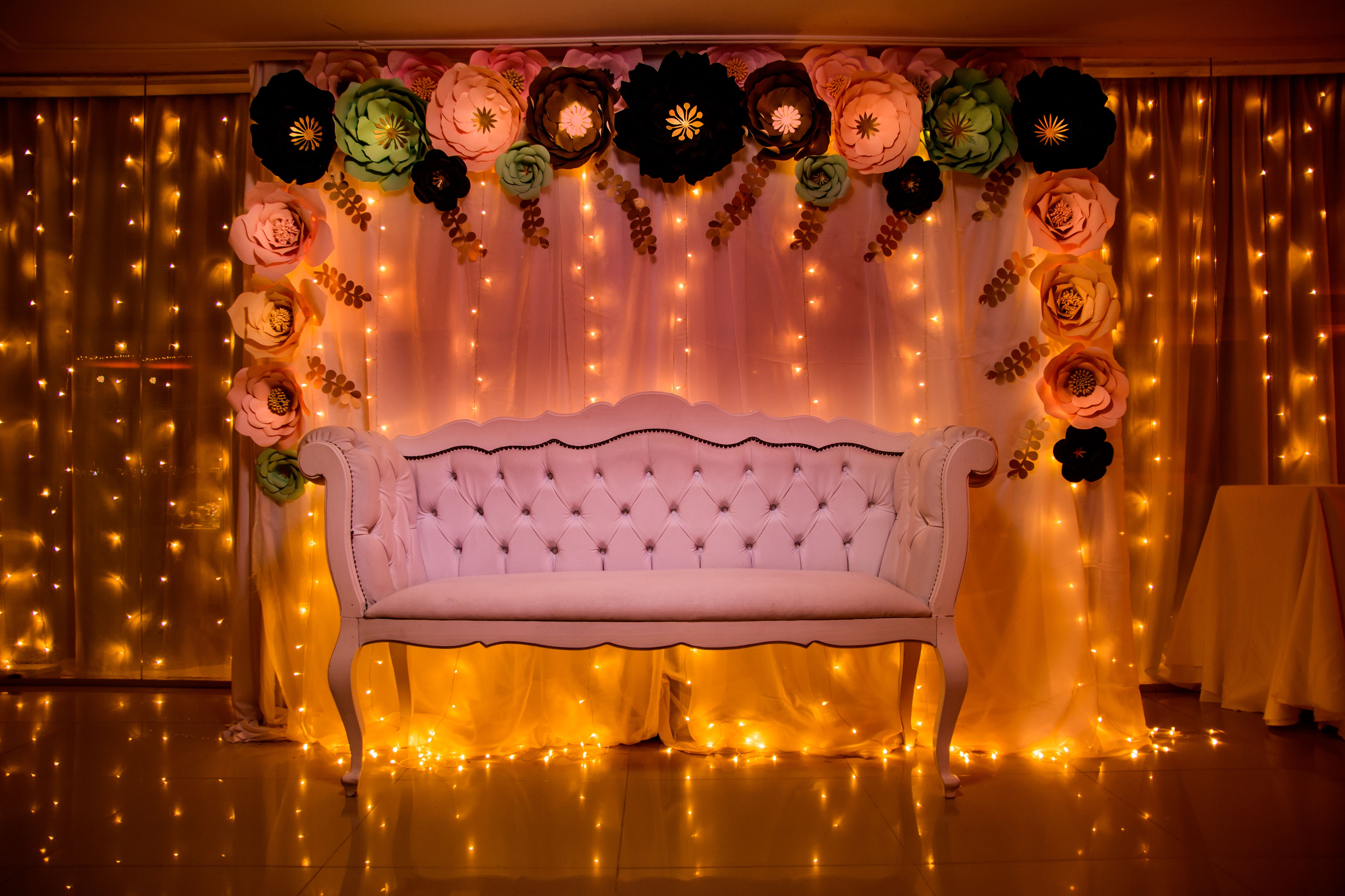 A beautifully decorated photo backdrop with a white sofa, paper flowers, and warm fairy lights.