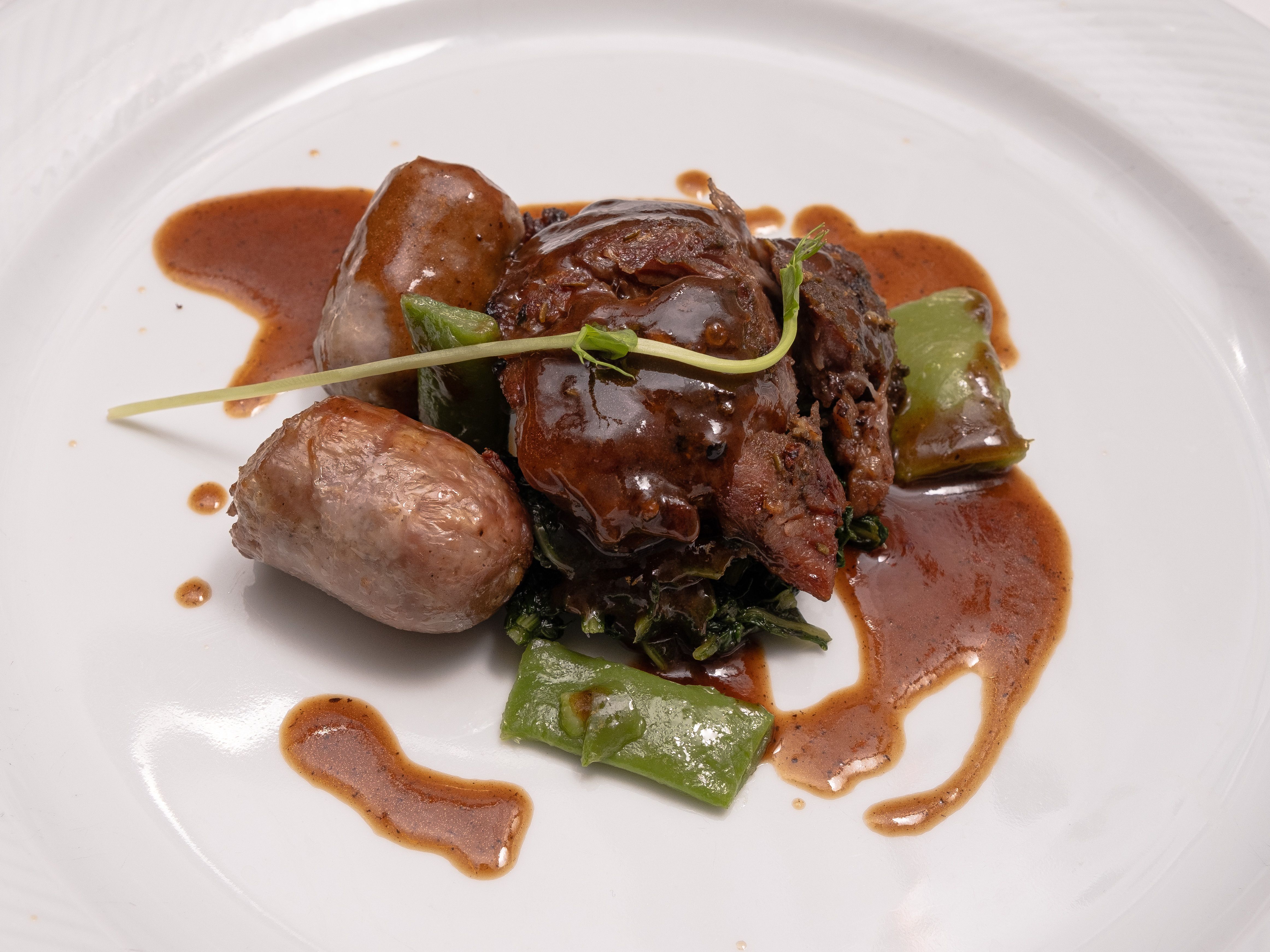 Gourmet beef dish served with sausage and greens on a white plate, elegantly presented.