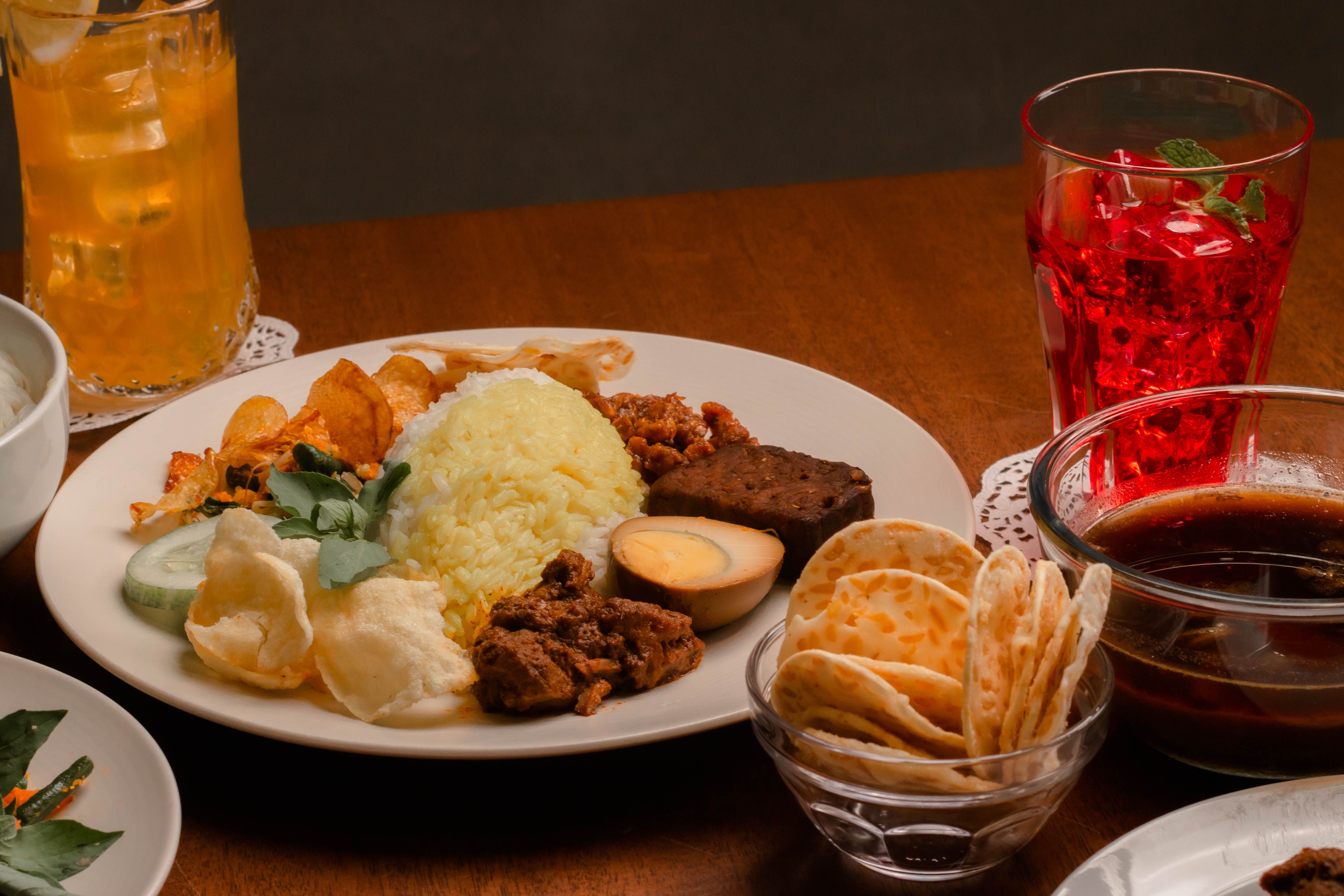 A delicious Indonesian meal including yellow rice, meats, crackers, and drinks served on a wooden table.