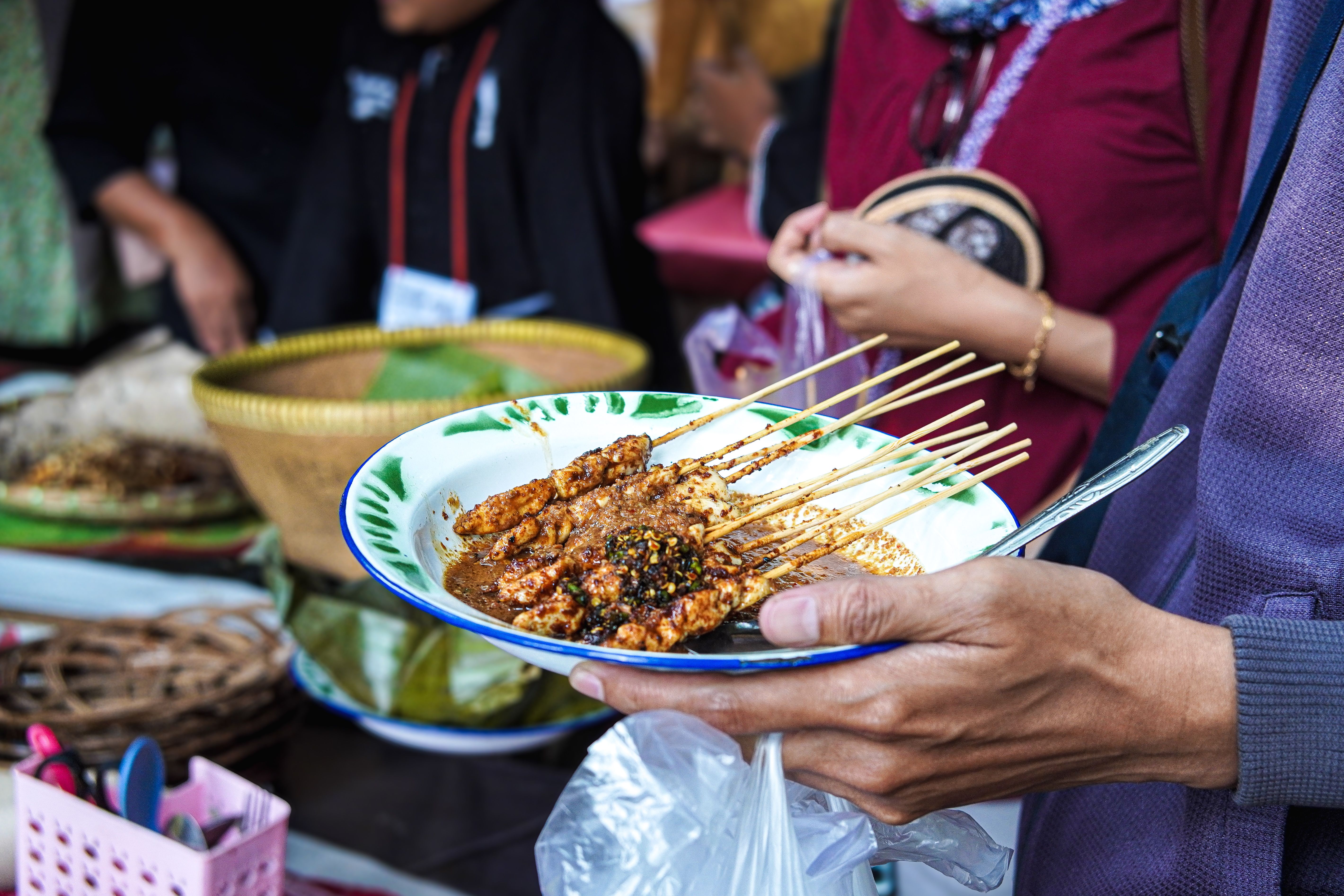 Close-up of a person holding a plate of Indonesian satay with peanut sauce at a street food stall in Purwakarta, Indonesia.