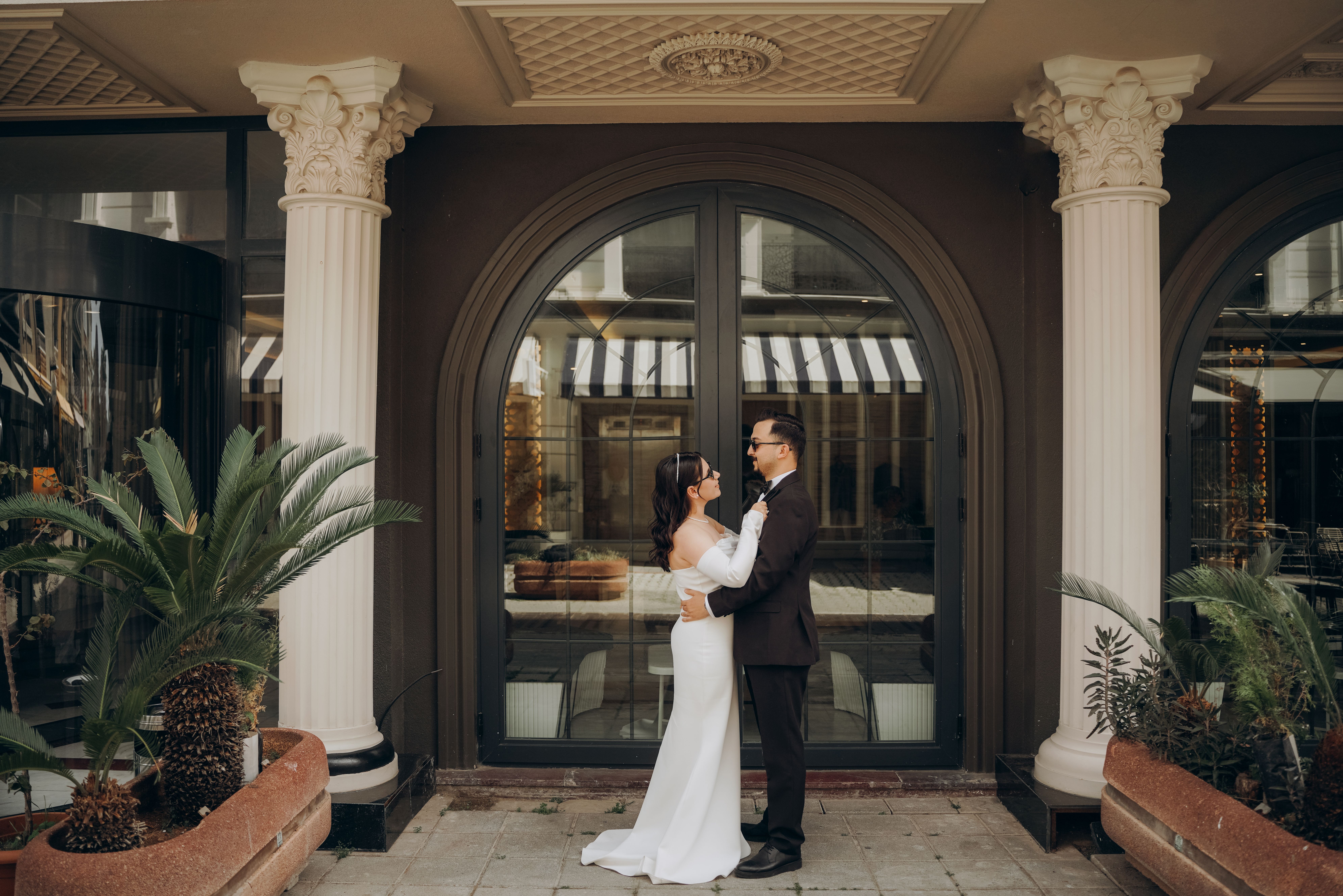 An elegant couple in wedding attire embracing outside a classic building with column details.